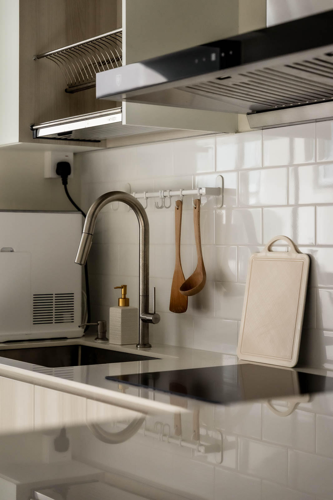 A modern kitchen with a stainless steel faucet, a soap dispenser, hanging wooden utensils, a cutting board, and an electric stovetop against a white tiled backsplash reminiscent of the sleek sophistication found at 36 Keppel Bay.
