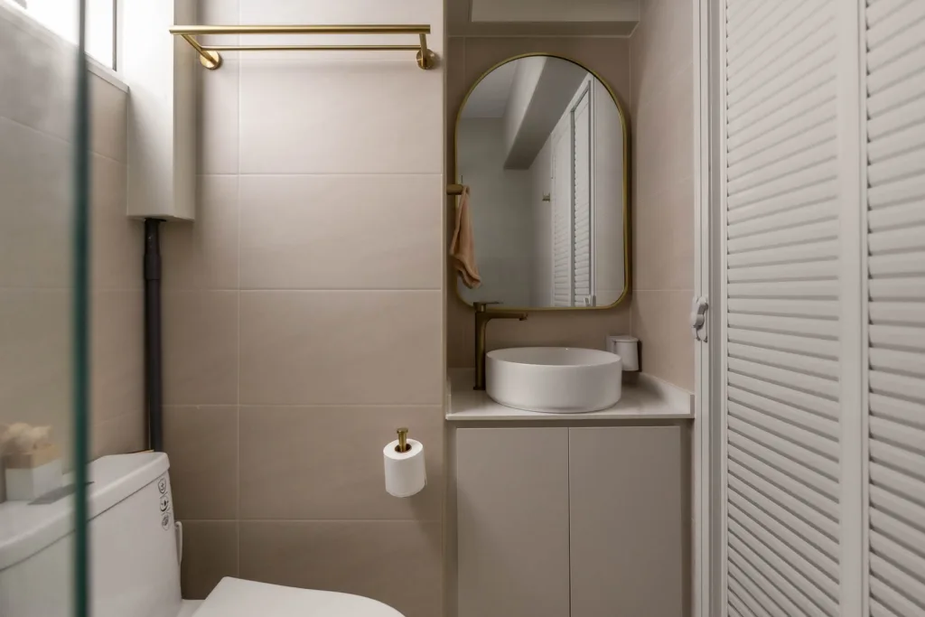 A compact bathroom featuring a white toilet, a wall-mounted sink with a round mirror above it, and a cabinet below. A towel rack and toilet paper holder are also visible, showcasing the sleek style often brought to life by Singapore interior design firms.