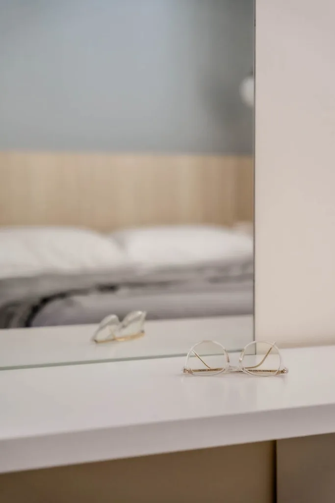 A pair of eyeglasses rests on a white surface in front of a mirror. In the reflection, influenced by loft interior design, a blurred bed with white pillows and sheets is visible.