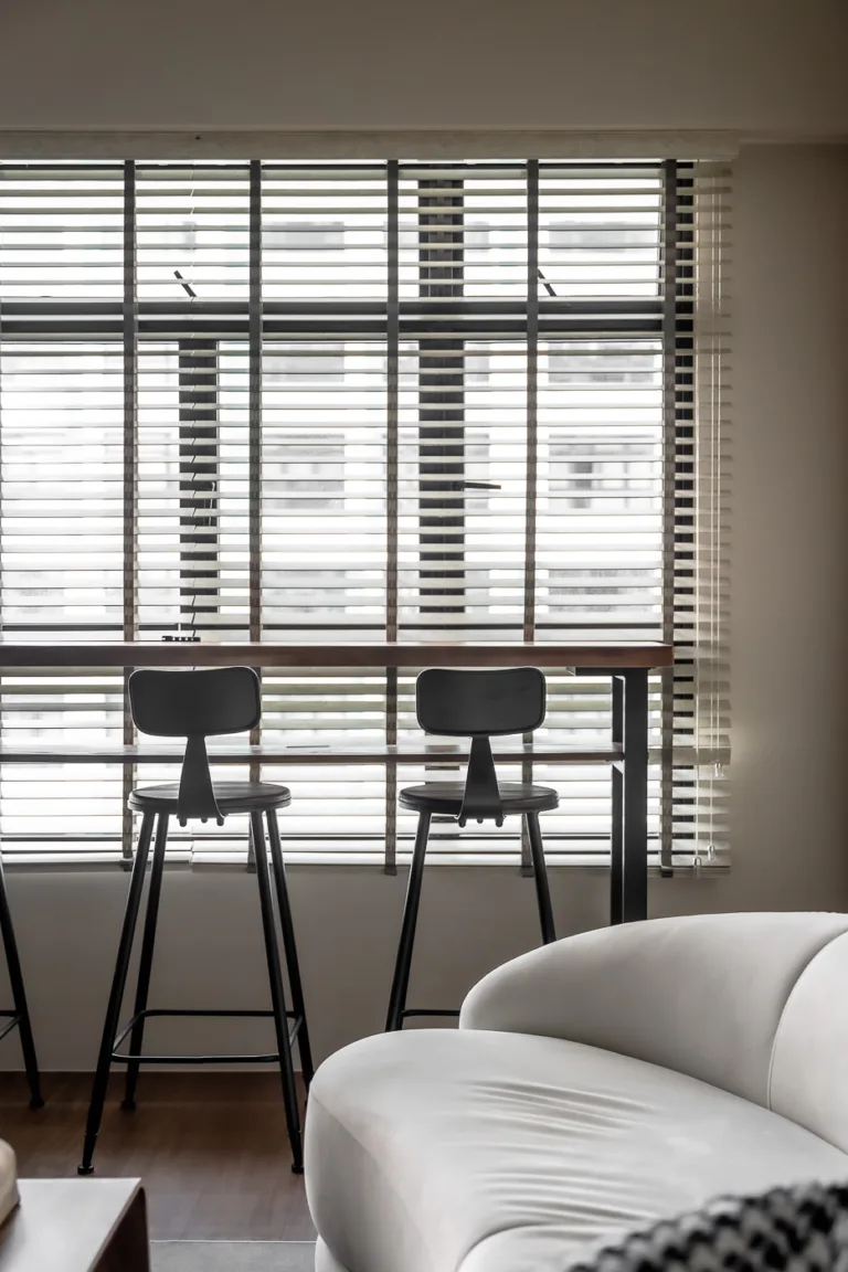 Modern living room with window blinds, wooden counter, black bar stools, and white curved sofa.