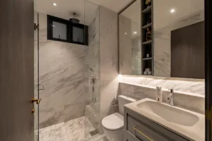 luxurious modern bathroom with marble walls and floor, glass shower enclosure, toilet, vanity with a sink, mirror, and built in shelves.