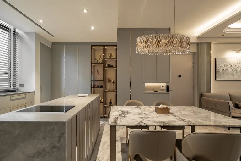 luxurious modern kitchen and dining area with marble countertops, an island, gray cabinets, a glass chandelier, and a dining table with beige chairs. decor includes small statues and a framed picture on the wall.